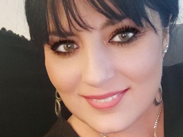 BrigitteXLove's face with dark brown eyes, black bangs, pink lipstick, and a small tattoo on her right cheek, wearing a black top and silver earrings.