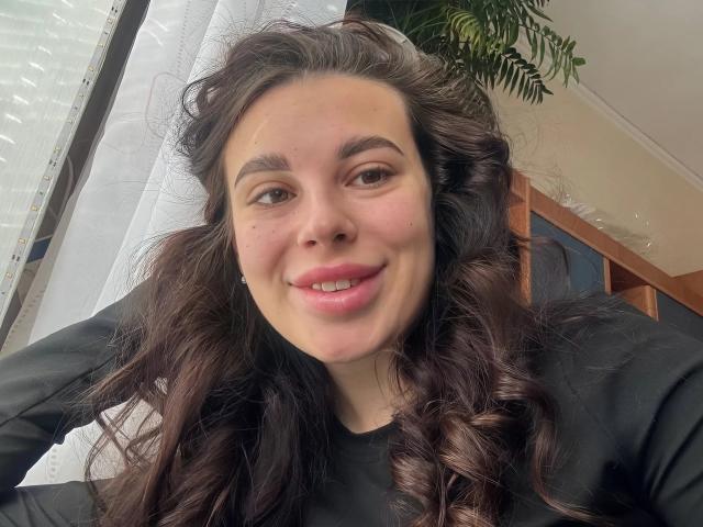 Portrait of BlackFatal, a young woman with wavy, dark brown hair, smiling slightly. She has light brown skin, full eyebrows, and a small mole above her right eye. She wears a black long-sleeve shirt. Her lips are pink and slightly parted. The background includes a white curtain and a potted green plant. The image has a natural, casual feel with soft, diffused light. Her hair is loosely curled.