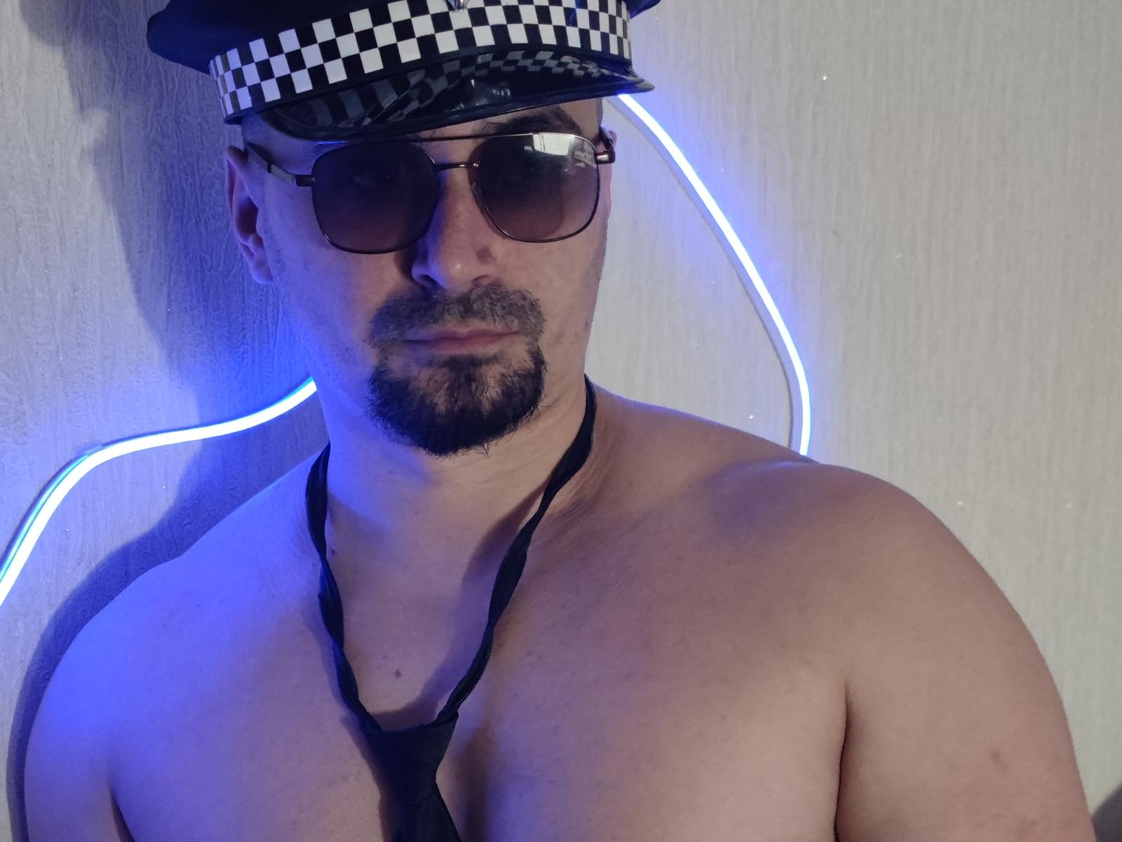 Ziesh is a shirtless man with a medium build and light skin, wearing a black and white checkered police cap and dark sunglasses. He has a goatee and is looking directly at the camera with a slight smirk. A black necktie hangs loosely around his neck. A blue neon light curves behind him, illuminating his upper body. His facial features are strong, and his chest is prominently displayed. The overall composition is simple, with a focus on Ziesh's upper body and facial expression.