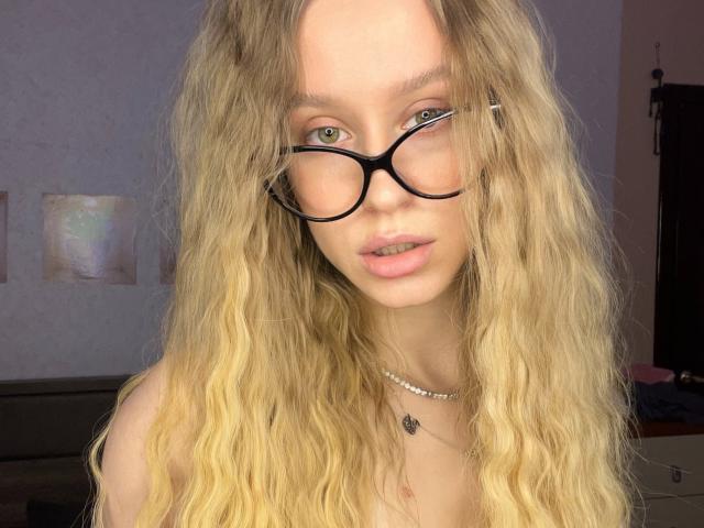 A close-up portrait of AliceGly. She has long, wavy blonde hair and wears black, thick-rimmed glasses. Her light green eyes are focused on the camera. She has a fair complexion and slightly parted pink lips. She wears a simple silver necklace with a small pendant. The background is blurry, with soft, warm lighting. The image has a natural, candid feel. Her hair is slightly messy, adding to the casual look. The focus is sharp on her face, with a slight blur on the background.