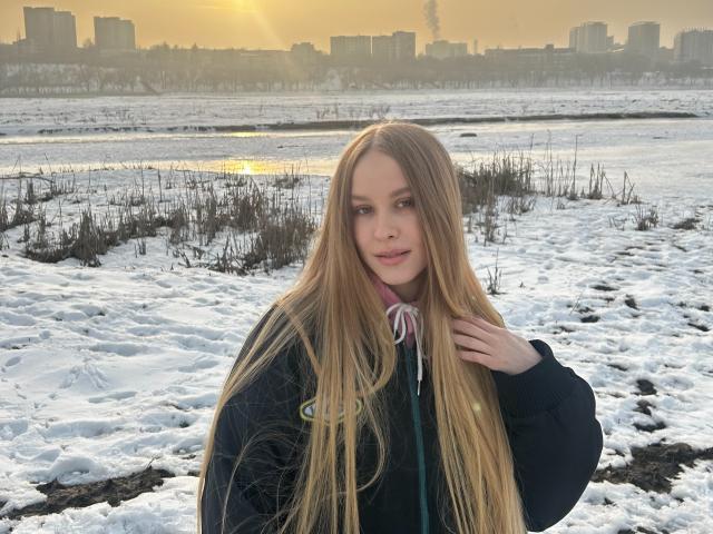 AliceGly stands in a snowy landscape, long blonde hair flowing past her shoulders. She wears a dark green hoodie with a pink ribbon at the collar. Her right hand touches her hair. The sun is low in the sky, casting a golden light. Background includes a distant city with tall buildings and a visible smokestack. Snow covers the ground, with patches of grass. AliceGly's expression is calm, with subtle pink lips.