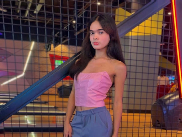 PeachAnderson-hot with long brown hair, pink strapless top showing small tits, and blue pants, standing in front of a metal grid, looking straight at the camera.