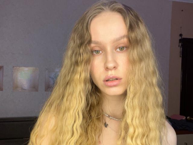 Close-up of AliceGly with long, wavy blonde hair, pale skin, and green eyes. She wears a small black necklace with a cross pendant. Her lips are slightly parted, and she looks directly at the camera. The background is blurred, with a dark object and light-colored wall. The lighting emphasizes her hair's texture and her facial features. Her expression is neutral. The image has a natural, slightly soft focus.