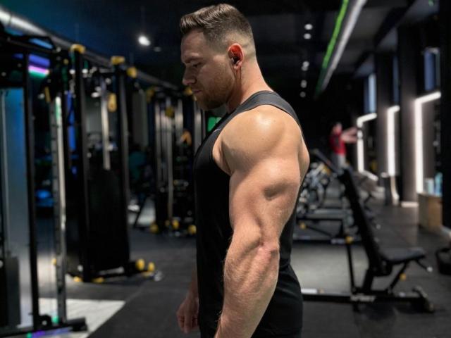 Thori69, ein muskulöser Mann mit einem kräftigen Arm, trägt ein schwarzes Tanktop, hat kurze, braune Haare und steckt Kopfhörer ein. Er steht in einem Fitnessstudio, blickt nach unten und spannt seine Muskeln an..