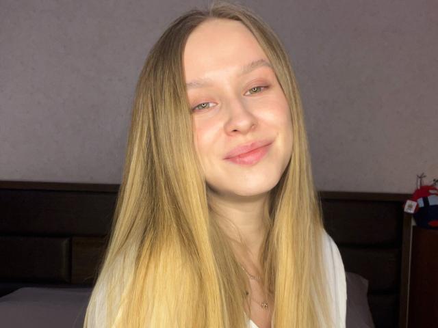 AliceGly is a young woman with long, straight blonde hair and fair skin. She has light green eyes and a subtle, content smile. Her pink lips are slightly parted. She wears a white shirt, and a thin necklace is visible. The image is a close-up portrait, focusing on her face and upper shoulders. The light source is behind her, creating soft shadows and highlighting her hair's smooth texture. The background is a simple, muted wall.