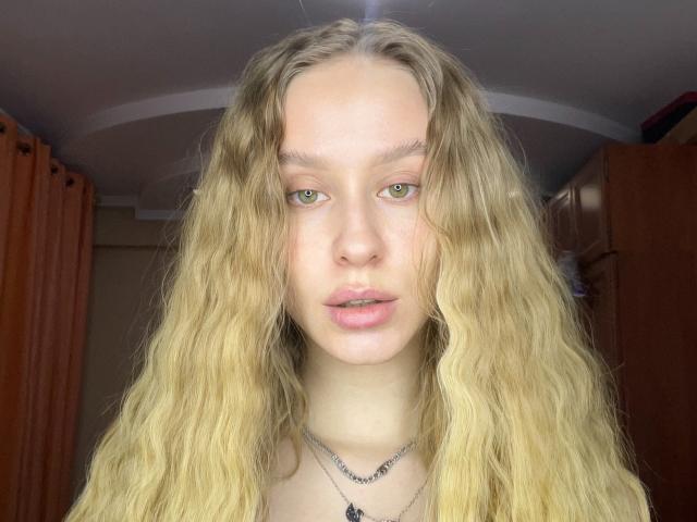 AliceGly has wavy blonde hair, green eyes, and a neutral expression. She's wearing a black necklace with a small pendant, and her pink lips are slightly parted.