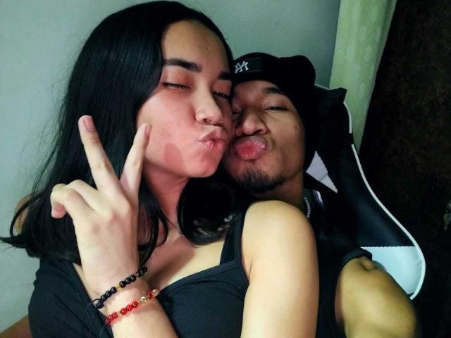 CoupleHotxz, with black hair and medium tits, makes a kissy face, holding up a finger, while CoupleHotxz, in a NY cap, sticks out tongue, sitting behind her.