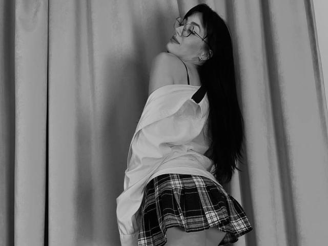 EviLune with glasses, long dark hair, off-shoulder shirt, plaid skirt, leaning back, showing her bare shoulder and black bra strap, looking up. Black-and-white photo.