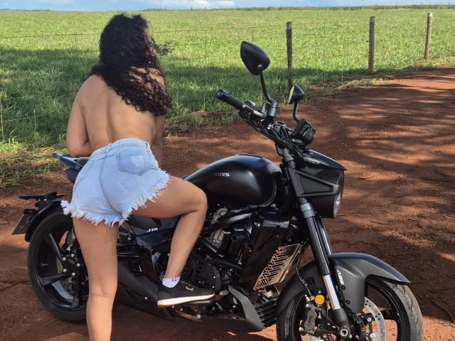 Nimety, topless with curly black hair, wearing frayed blue shorts and black shoes, sits on a black motorcycle, showing her ass, on a red dirt road.