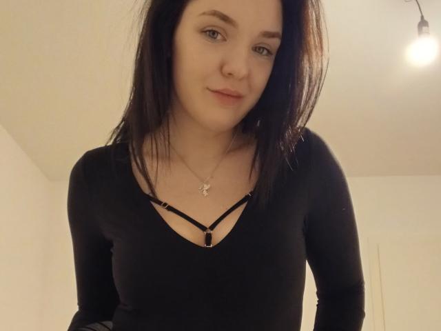 MrsXSweet with black hair, wearing a black V-neck top with strappy details, showing her tits, and a necklace with a small pendant, smiling slightly.