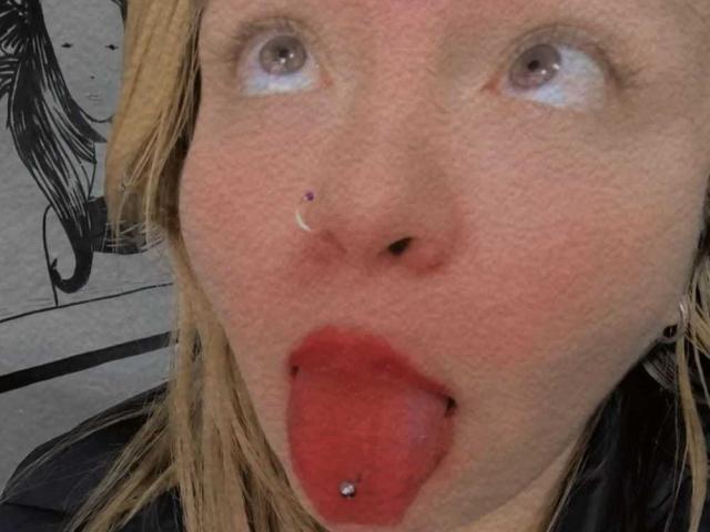 SophieeBanks has a nose piercing, red lips, and a tongue piercing with a ball on it. Her blonde hair is down, and she's sticking her tongue out.