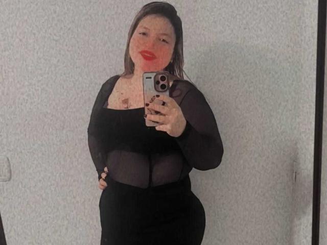 SophieeBanks with red lipstick, black sheer top, and black skirt, holding phone, showing her curvy, busty figure, and plump ass, in a mirror selfie.