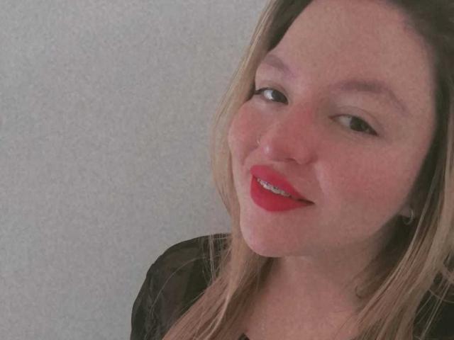 SophieeBanks has red lipstick, a nose piercing, and a slight smile with her head tilted. She's wearing a black top with blonde hair falling to her shoulders.