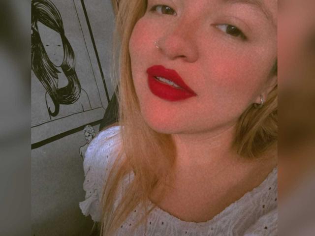 SophieeBanks with red lipstick, blonde hair, nose ring, and white shirt, looking at camera, soft pink blush on cheeks, black and white drawing of woman in background.