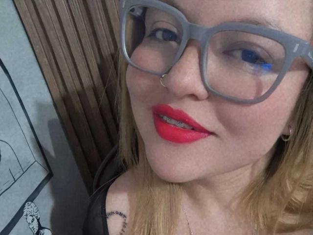 SophieeBanks with grey glasses, red lipstick, nose ring, and blonde hair, showing her teeth, wearing a black top, and a necklace with a small pendant.