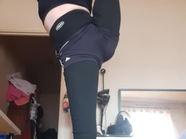 IssahneMay's black leggings are pulled up to her waist, exposing her bare lower belly and black sports bra, with one leg raised high. Her black underwear is partially visible.