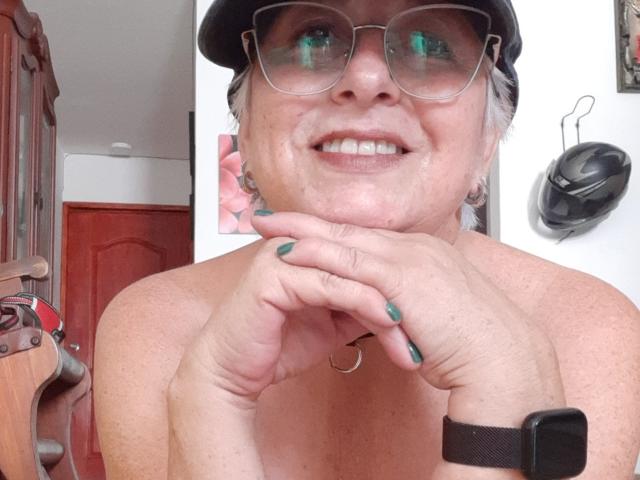 Canitas69 with gray hair, glasses, black cap, green nail polish, smiling, topless, hands near face, wearing black wristwatch, showing small, saggy tits, and a slight double chin.