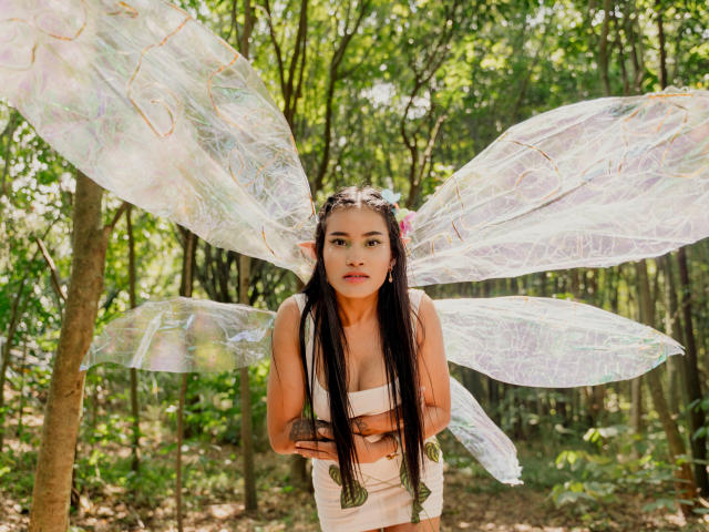 KarolPalacios with translucent butterfly wings, long black hair, wearing a white dress, standing in a forest, hands near chest, looking forward, elf-like ears, green forest background.