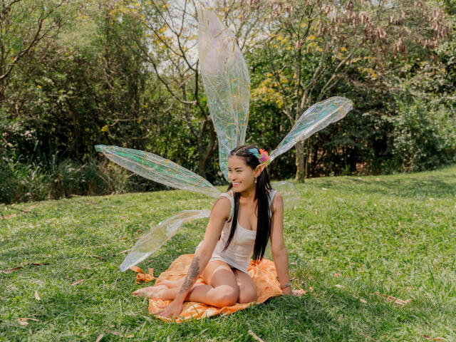KarolPalacios sitting on grass with transparent fairy wings, orange skirt, and white tank top, smiling, brown hair, tattoo on left forearm, flower in hair, sunlit, green background.