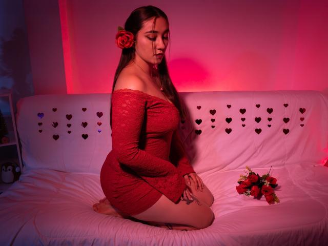 SwettieSammy kneeling on a white couch in a red lace off-shoulder dress, with a red rose in her hair, hands on her ass, pink and red lighting.