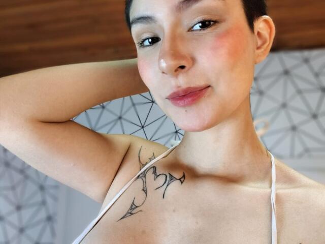 IsabellaHallad with short dark hair, light skin, small tattoos on her chest, and a slight smile, wearing a white halter top, arms raised, showing her small tits.