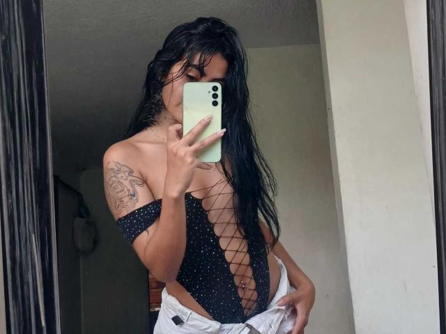 NicolleSweetty with wet black hair, wearing a black, polka-dot, fishnet, one-piece, exposing her tits and a tattoo on her right arm, taking a mirror selfie.