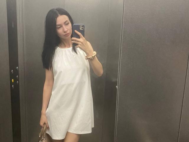 StarrySiren with long black hair, wearing a white sleeveless dress, holding a phone, taking a mirror selfie in a dark elevator, with a beige bag in her left hand.