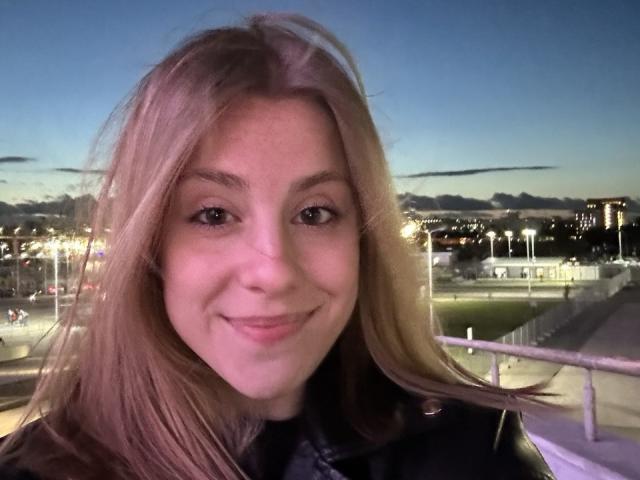 KerryColver's smiling face, blonde hair, and light skin, wearing a black leather jacket, with small tits, in a selfie at dusk, city lights in the background.