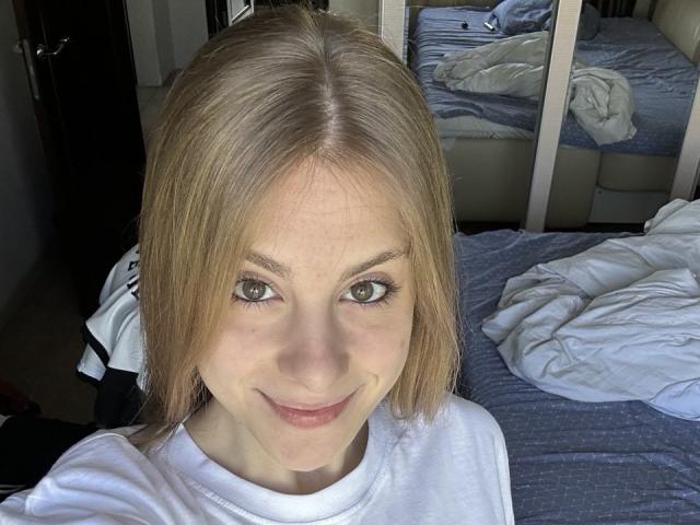 KerryColver with blonde hair, green eyes, and a slight smile, wearing a white shirt, in a bedroom with a messy bed and mirror. Her ass is not visible.