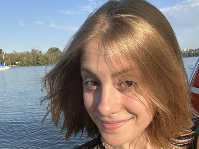 KerryColver's face with light brown, wavy hair, smiling subtly. She has fair skin, green eyes, and a slight pink nose. Background shows a calm blue waterway and trees.