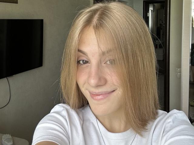 KerryColver has blonde, straight hair covering one eye, a slight smile, and is wearing a white shirt with a silver necklace. Her tits are not visible.