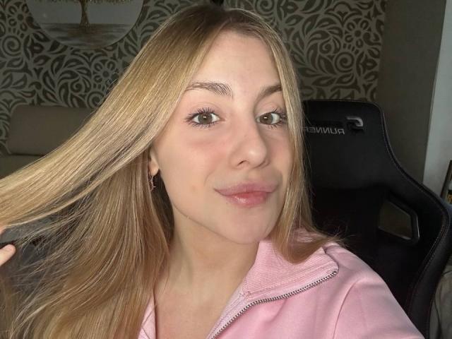 KerryColver with blonde hair, light skin, and pink lips, wearing a pink zip-up, holding her hair, sitting in a black chair, looking at the camera, smiling softly.