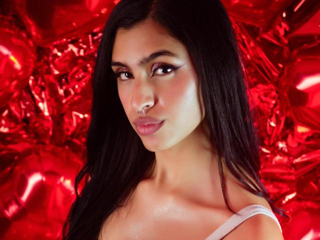 LilithDesire has long black hair, brown eyes, and a nose ring. She's wearing a white top, with shiny skin and a slight smile, set against a red, crumpled background.