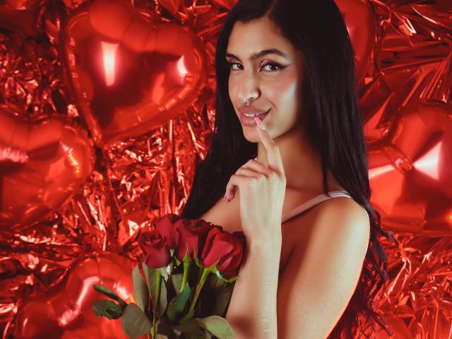 LilithDesire with long black hair, holding red roses, finger to lips, smiling, wearing a strapless top, red heart balloons in background, looking sexy and cute.