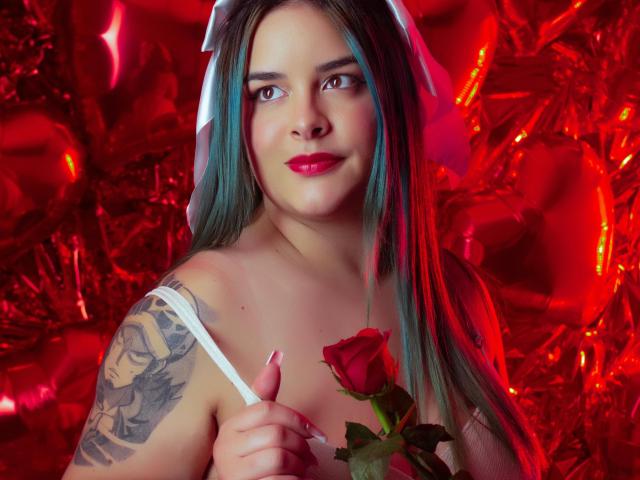 AuroraSantorini com cabelo azul-turquesa, batom vermelho, alça branca, segurando uma rosa na mão, pressionando-a contra os seios, tatuagem no braço esquerdo, sorrindo, fundo vermelho, lenço branco na cabeça..
