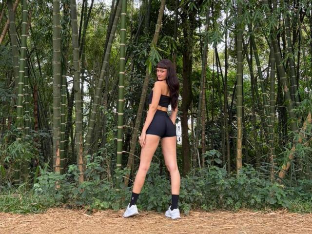 CandyyClair stands in a forest, facing away, wearing a black sports bra, black shorts, black socks, and white sneakers, holding a white water bottle, showing her ass.