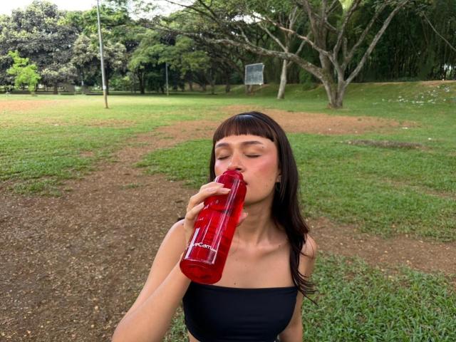 CandyyClair with black strapless top, drinking from a red water bottle, eyes closed, brown hair with bangs, green grass and trees in the background, holding bottle in right hand.
