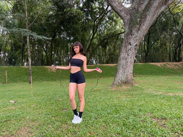 CandyyClair with long brown hair, black strapless top, black shorts, white sneakers, jumping rope in a grassy park, holding pink handles, tree in background, green trees behind.