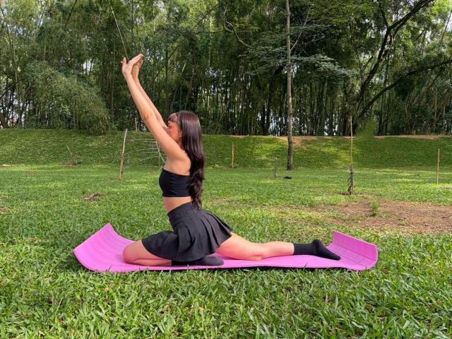 CandyyClair, black sports bra, black skirt, pink yoga mat, stretching, arms raised, front split, green grass, forest, long dark hair, visible butt, socks on.