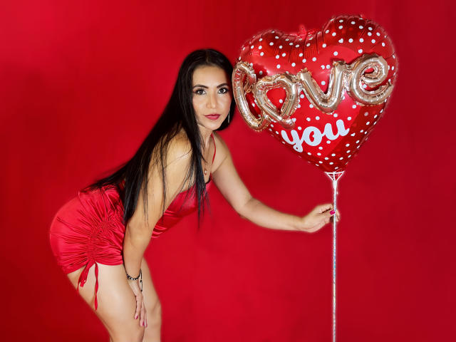 VivienneWest with long black hair, wearing a red satin dress, holding a heart-shaped balloon with "love you" and white polka dots, bent forward, showing her ass.