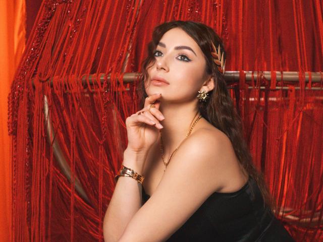OliviaCluff has wavy brown hair, golden earrings, and a black top, resting her chin on her hand with a subtle smile. Red beads and gold bracelet.