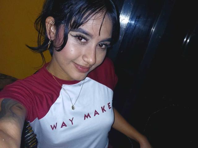 NicolleSweetty with dark hair, hoop earrings, and a red-white rag tee, smiling at the camera, showing small tits, a tattoo on her arm, and a gold necklace.