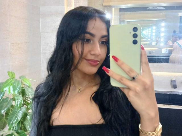 NicolleSweetty with long black hair, red lipstick, and red nails, holding a green phone, wearing a black top, taking a mirror selfie, showing off her boobs.