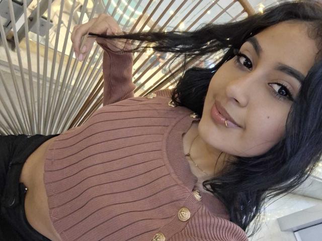 NicolleSweetty with long black hair, holding a strand, wearing a ribbed pink top with gold buttons, smiling at the camera, showing her small tits and belly button.