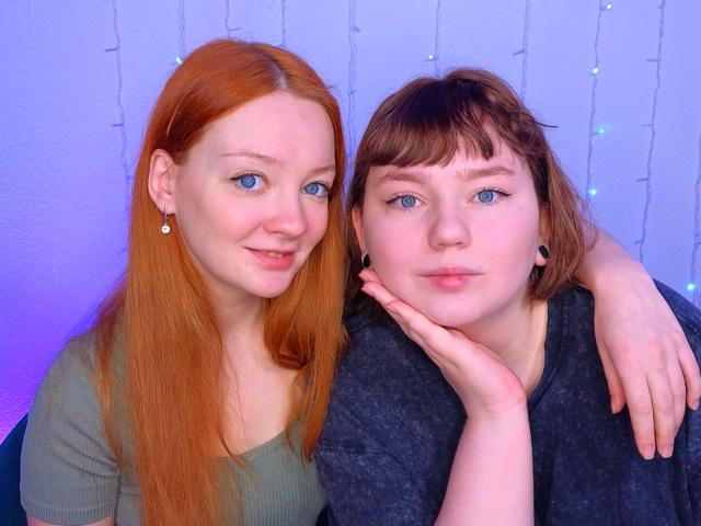 Two RadiantStwo's with red and brown hair, blue eyes, and fair skin, wearing green and gray shirts, sitting close, one with hand on cheek, smiling, in soft blue light.
