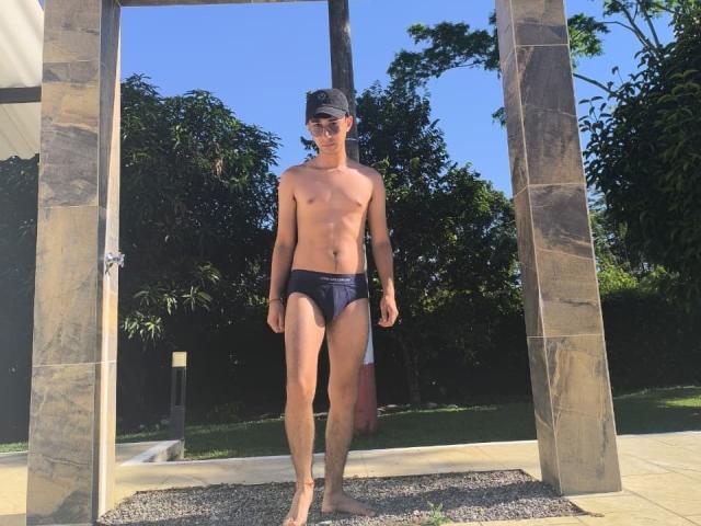 OliverHot69, ohne Hemd, trägt dunkelblaue Unterwäsche, eine schwarze Kappe und eine Sonnenbrille, steht in einem von der Sonne beschienenen Steingestell, mit blauem Himmel und grünen Bäumen im Hintergrund..