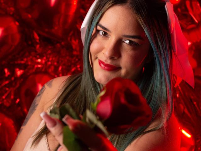 AuroraSantorini with green-tinted hair, white headpiece, red lipstick, holding a red rose, smiling softly, tattoos on her arm, red illuminated background, photo with a sensual vibe.