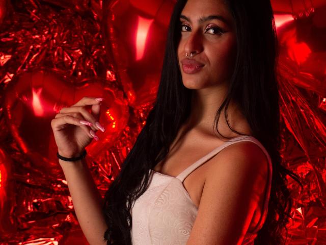 LilithDesire with long black hair, nose ring, and pink nails, wearing a white lace dress, holding her hand up, red lighting, and a sly smile on her face.