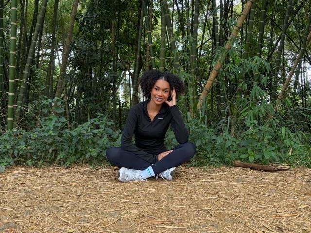 AylaCallowey with curly black hair, smiling, sitting cross-legged on pine needles, wearing a black long-sleeve shirt, black pants, and blue sneakers, in a bamboo forest.