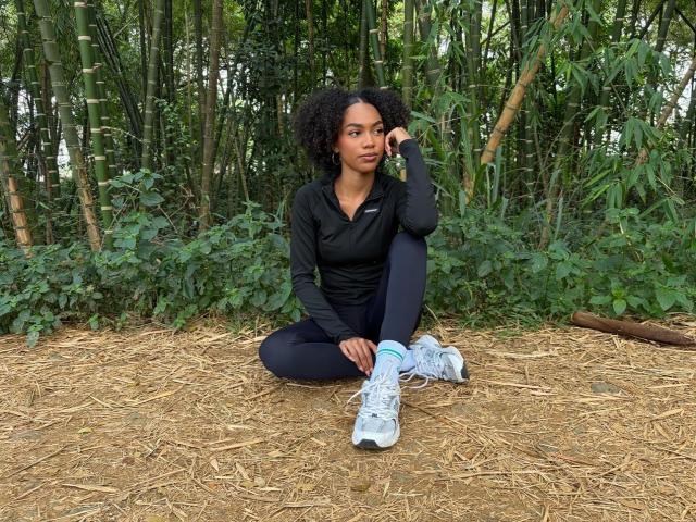 AylaCallowey, with curly black hair, sits on a dirt ground in black workout clothes and white sneakers, hand on chin, looking serious, surrounded by green bamboo.